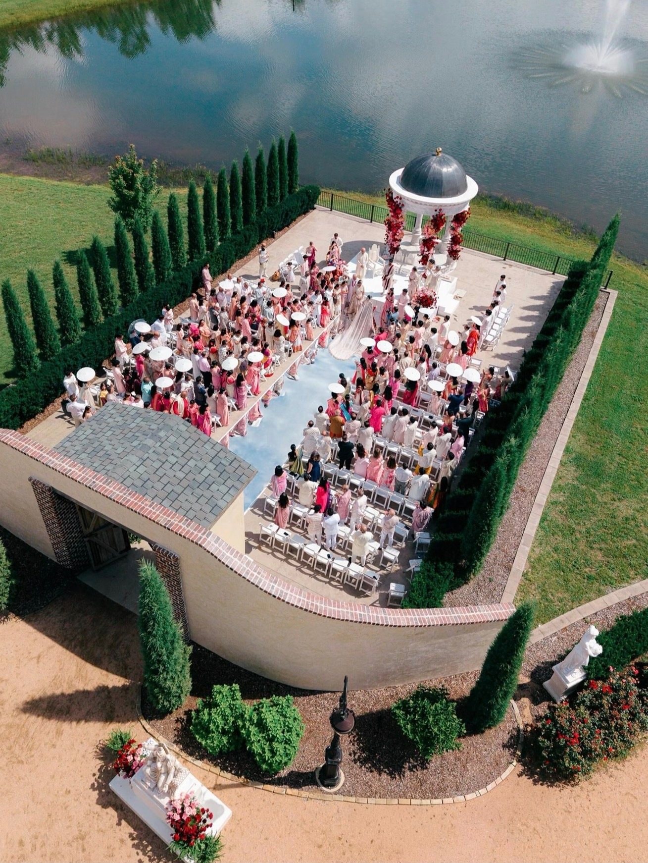 An aerial view of an outdoor ceremony with guests in colorful attire seated before a white gazebo, beside a pond with a fountain.