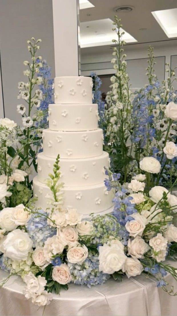 A tall white tiered cake surrounded by white roses, hydrangea and blue delphinium.