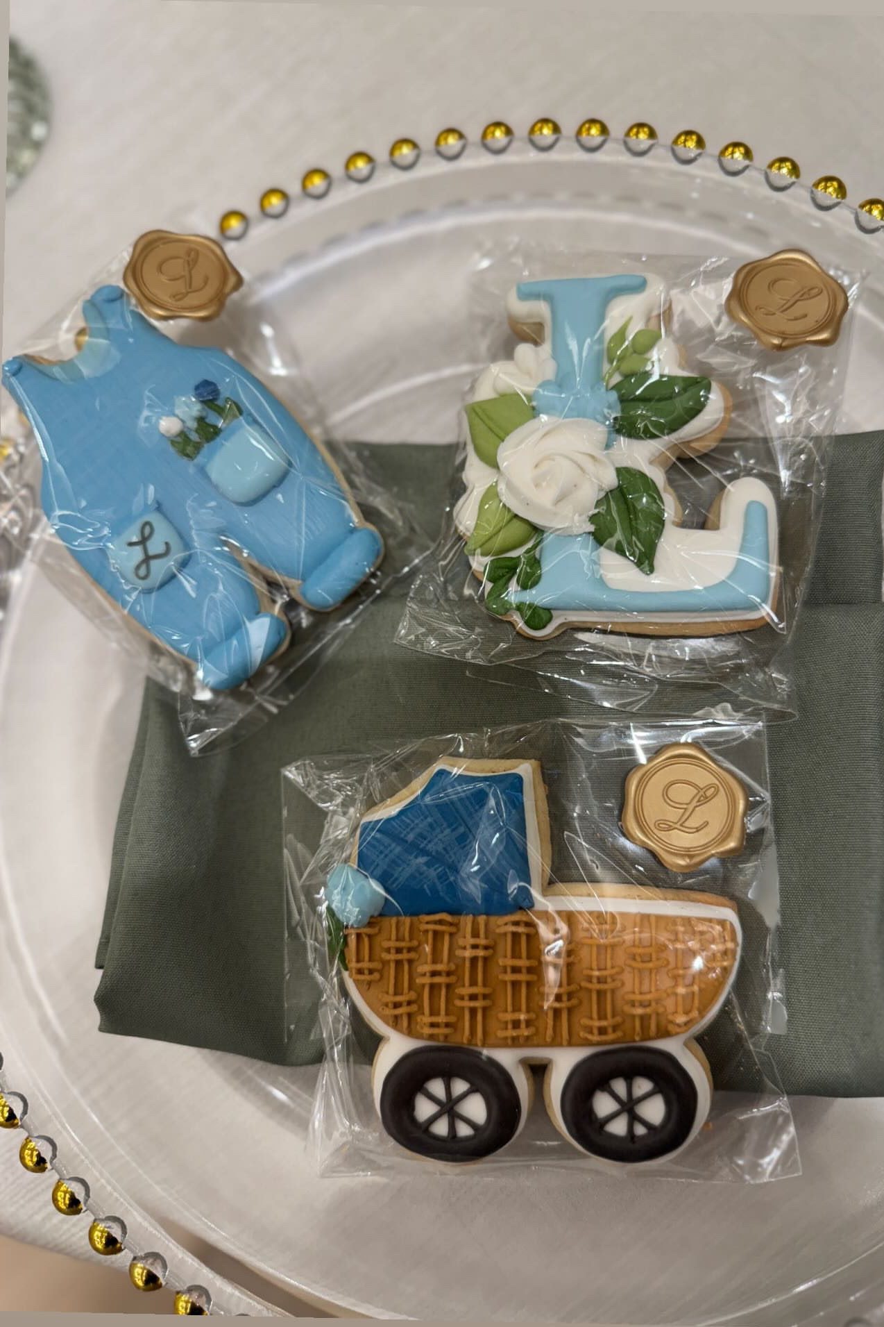 Individually wrapped decorated cookies shaped like a blue baby romper, a floral letter L and a woven baby carriage, each sealed with a gold monogram sticker, on a gold-beaded charger.