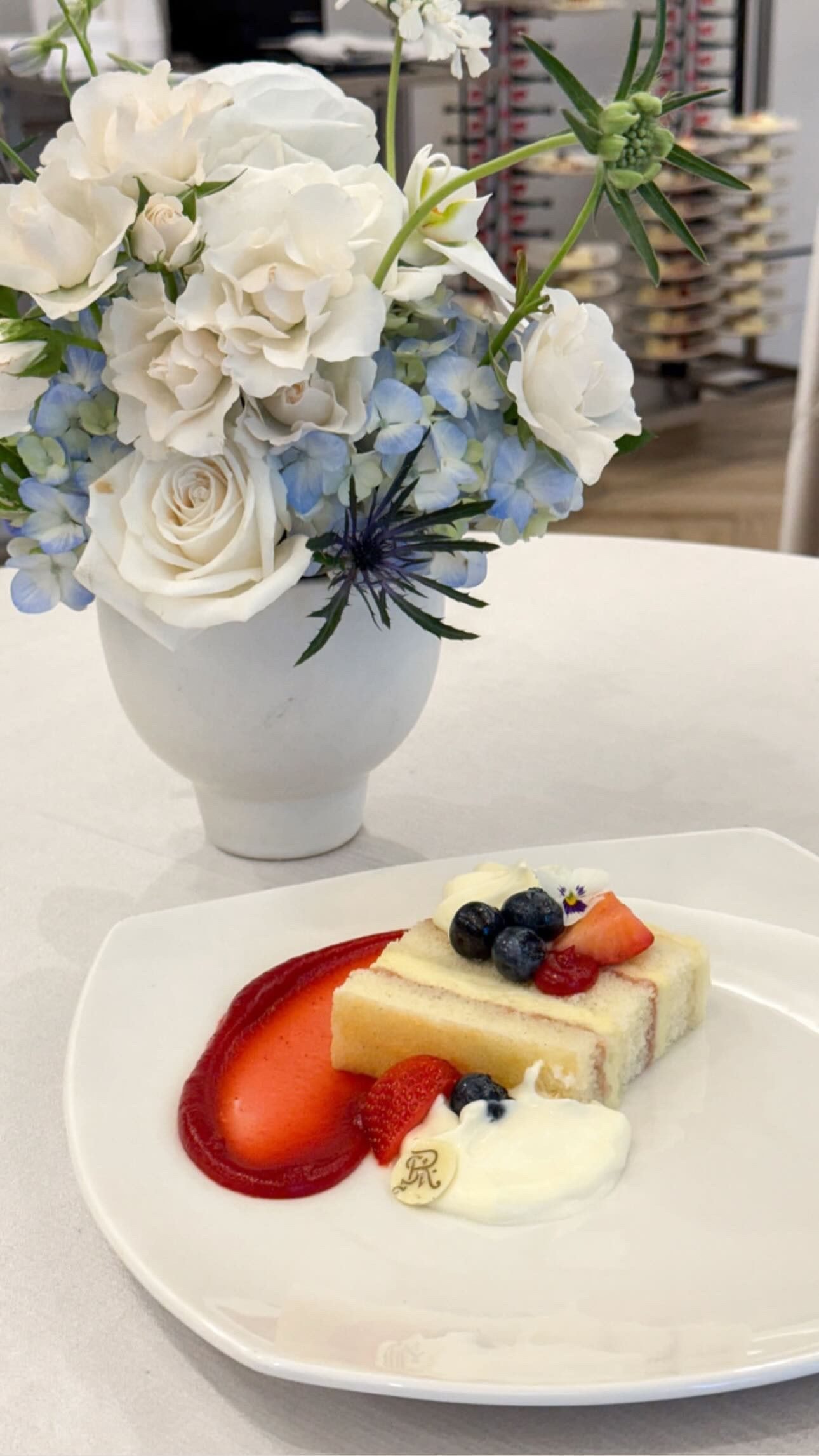 A slice of layered sponge cake with berries and strawberry sauce on a white plate, beside a vase of white roses, blue hydrangea and blue thistle.