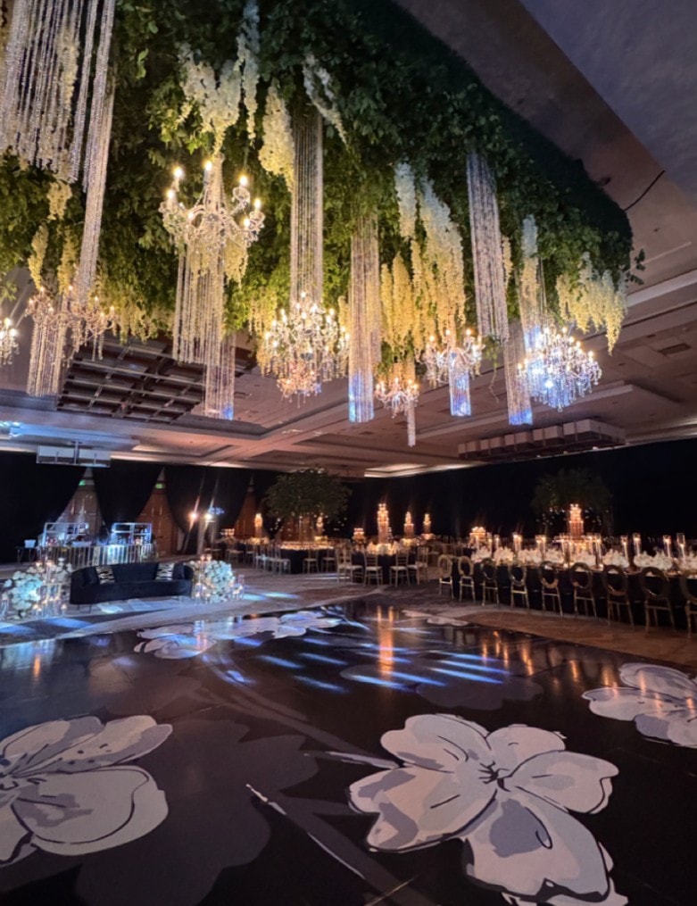A ballroom set for a celebration, with hanging greenery, white wisteria and crystal chandeliers above a dark dance floor painted with large white flowers, and long candlelit tables.