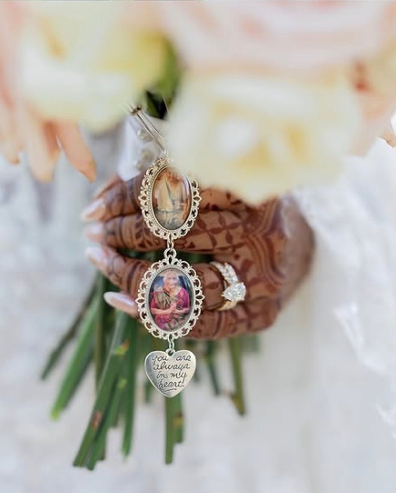 A bride’s mehndi-covered hand holds a bouquet wrapped with two framed photo charms and a heart-shaped charm.