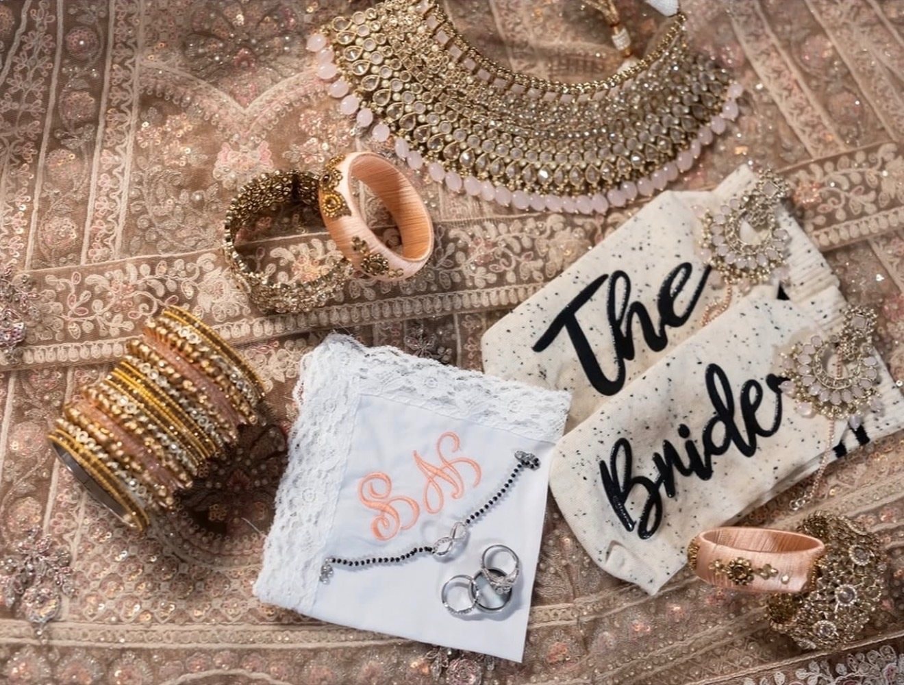 A flat lay of bridal jewelry, bangles and rings with a hanger tag reading ‘The Bride’ and a monogrammed handkerchief on embroidered fabric.