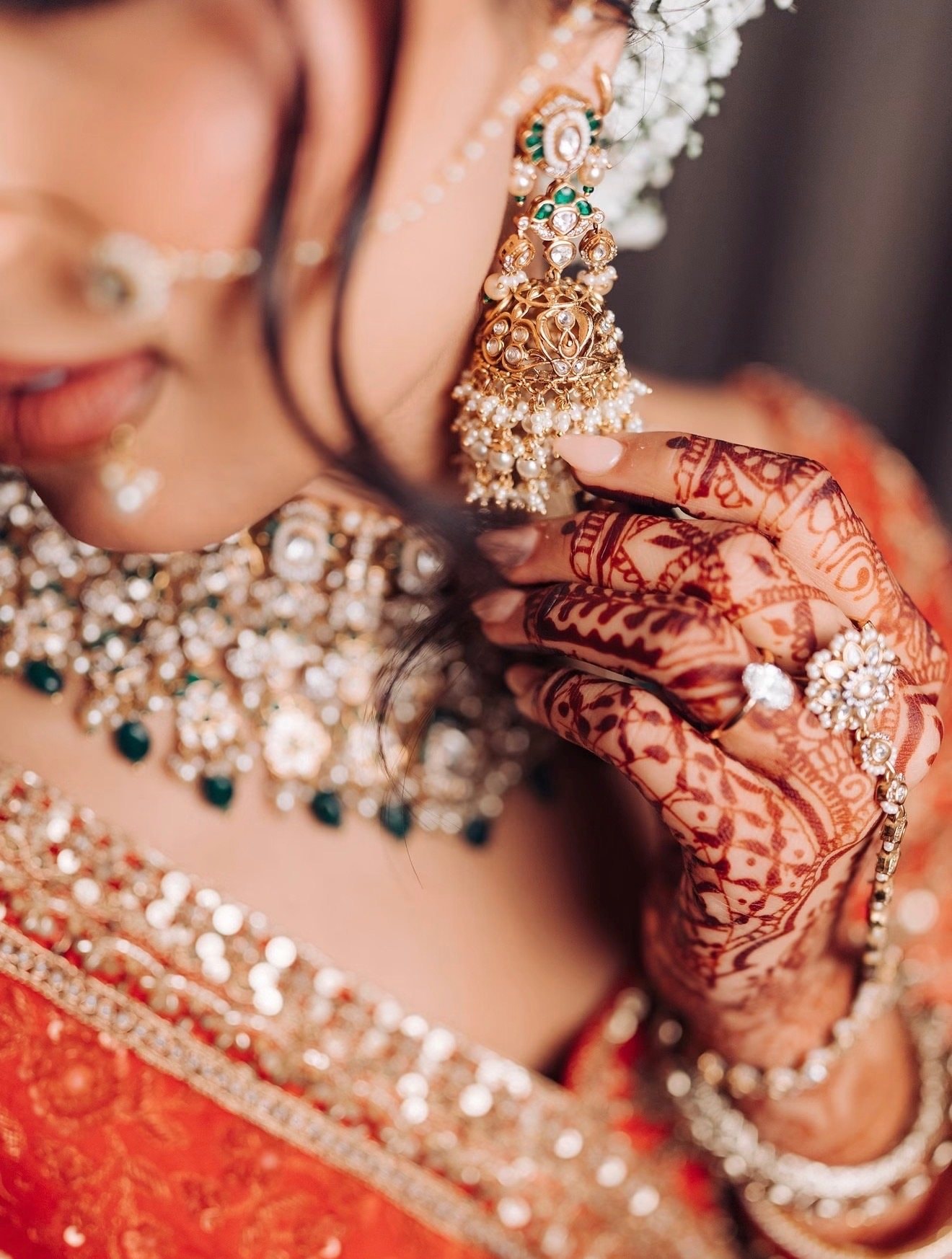 Close-up of a bride’s kundan earring, emerald and pearl choker, mehndi-patterned hand and rings against a red outfit.