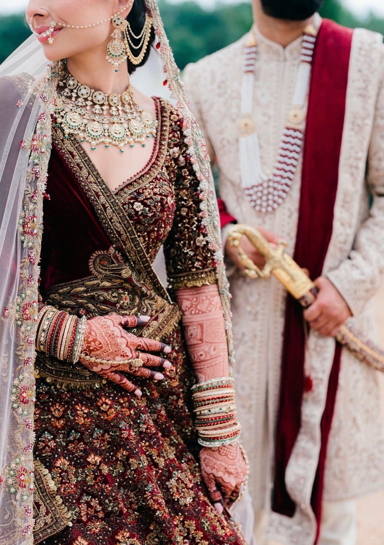 A bride in a maroon embroidered lehenga with a nath and kundan jewelry stands beside a groom in an ivory sherwani with a maroon dupatta, his hand on a ceremonial sword.