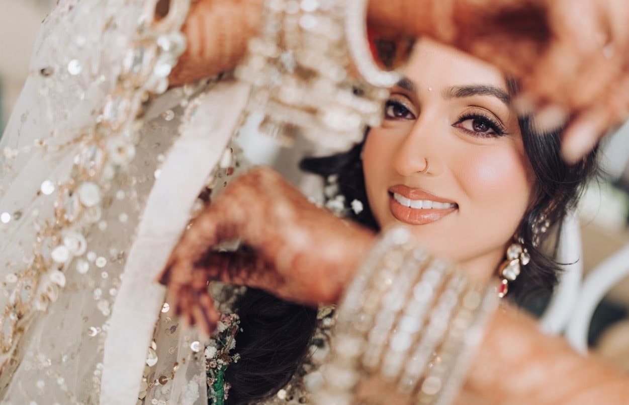 A smiling bride frames her face with her mehndi-covered hands and stacked bangles, a sequined ivory dupatta over her hair.