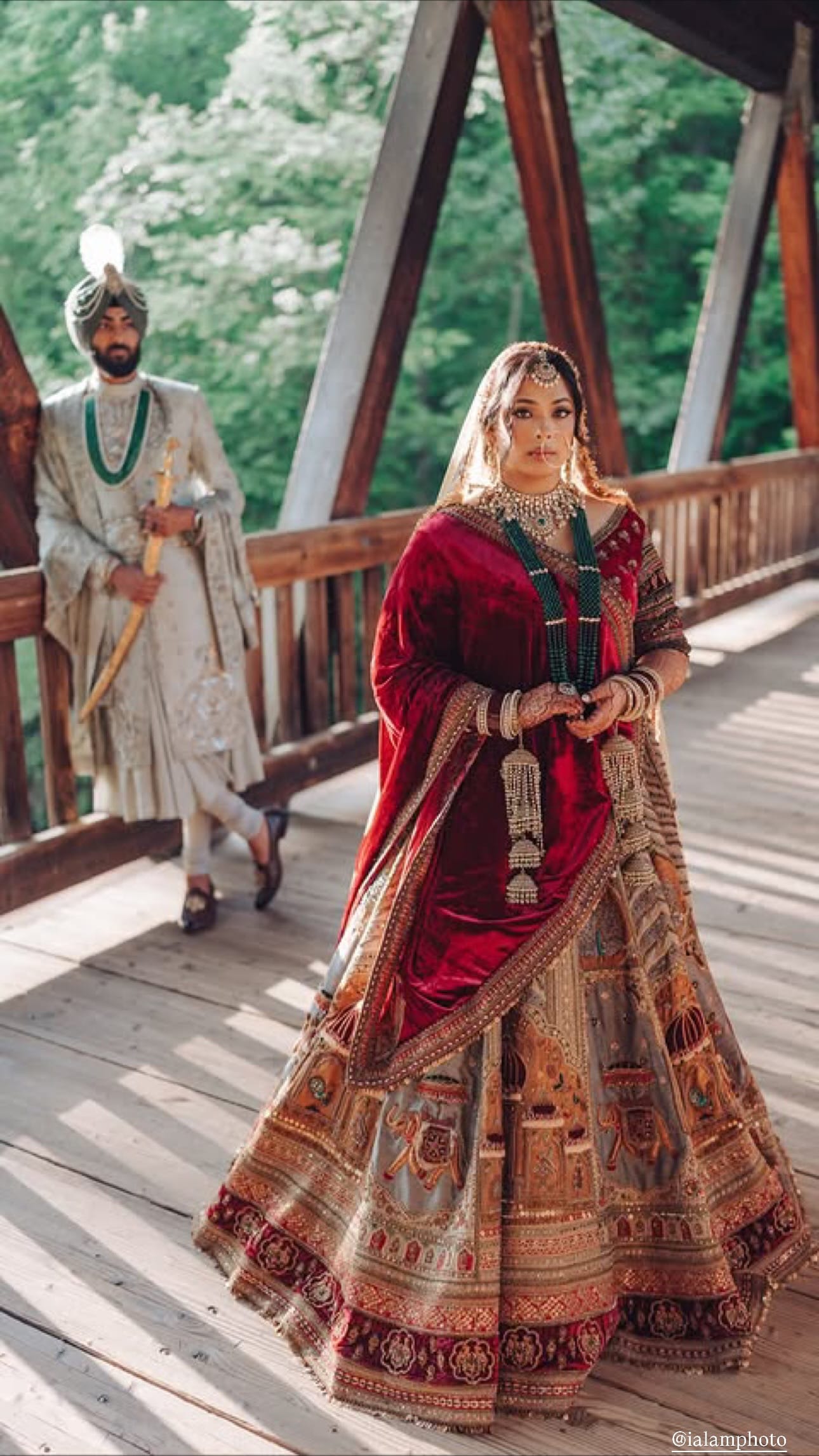 A bride in a red velvet dupatta and a hand-painted lehenga with elephant and palace motifs walks across a wooden covered bridge while a groom in a pale sherwani and turban waits behind her holding a ceremonial sword.