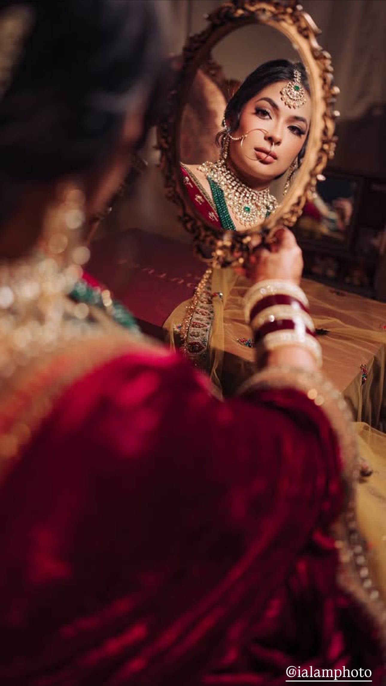 A bride in a deep red velvet dupatta and layered emerald and pearl jewelry studies her reflection in an ornate gold hand mirror, her bangled wrist raised.