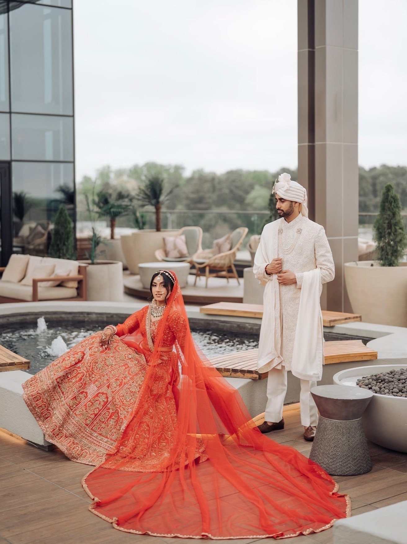 A bride in an orange embroidered lehenga with a long sheer dupatta sits on a rooftop terrace beside a fountain while the groom, in an ivory sherwani and turban, stands behind her.