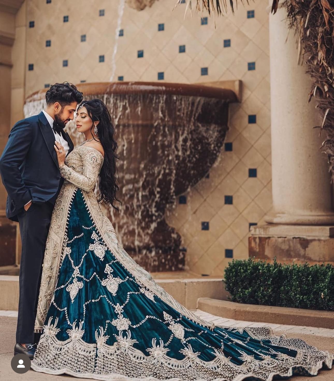 A bride in a teal velvet lehenga with silver embroidery and a long train leans into a groom in a navy tuxedo in front of a tiled stone fountain.
