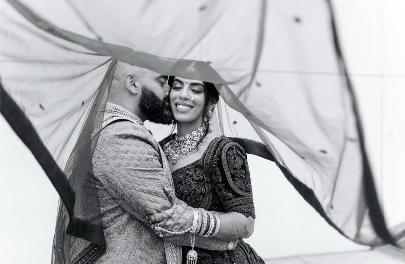 A black-and-white photo of a couple embracing as a sheer veil blows around them.
