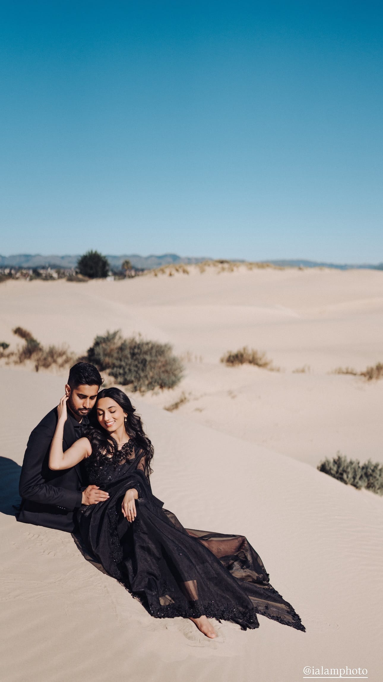 A couple in black formalwear, the woman in a sheer black embroidered sari, sits together on pale sand dunes under a clear blue sky.