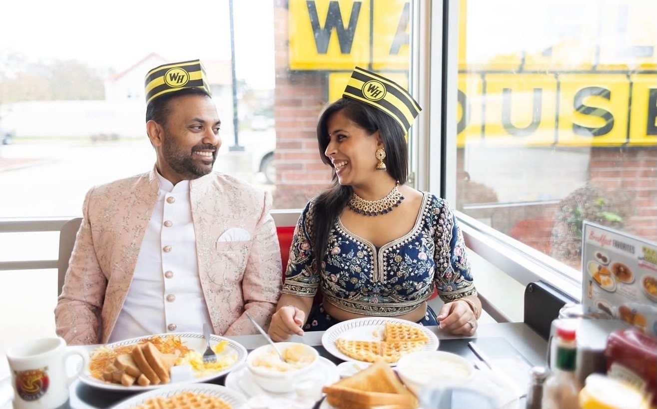 A couple in wedding attire and paper diner hats smile at each other over a table of waffles and coffee.