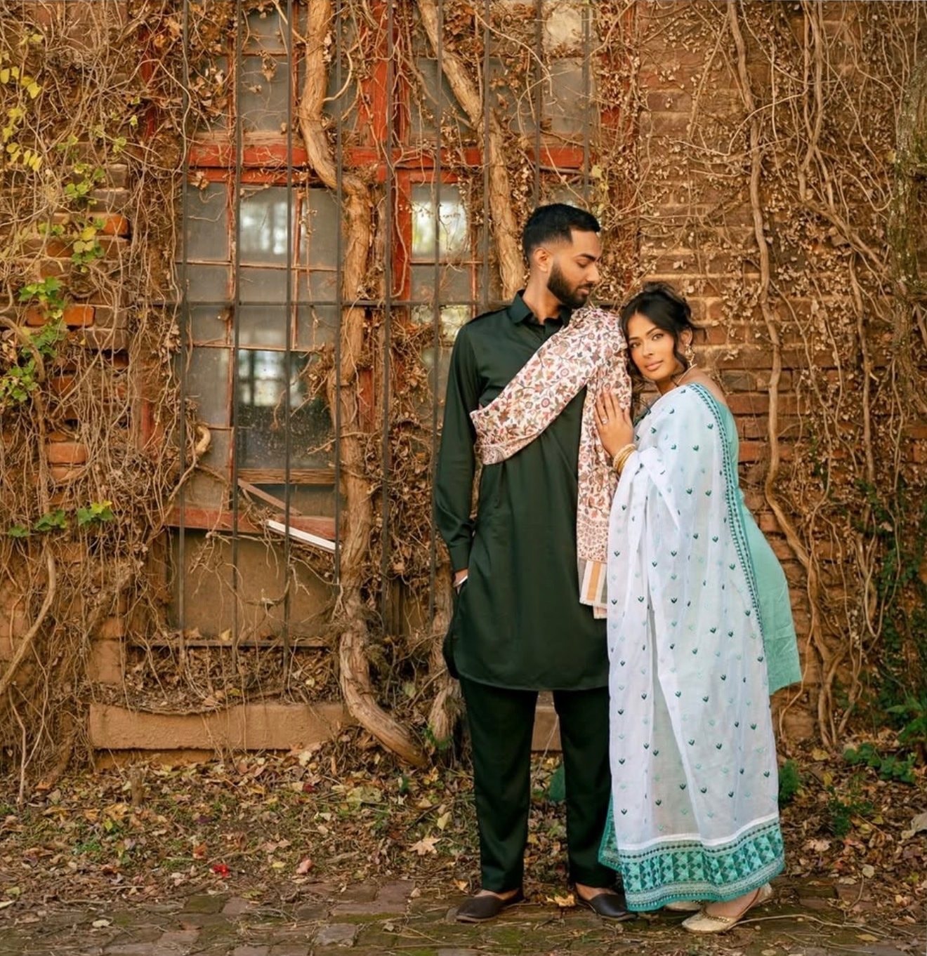 A couple stands before an ivy-covered brick wall, the man in a green kurta with a patterned shawl and the woman in a white and teal outfit with a dupatta.