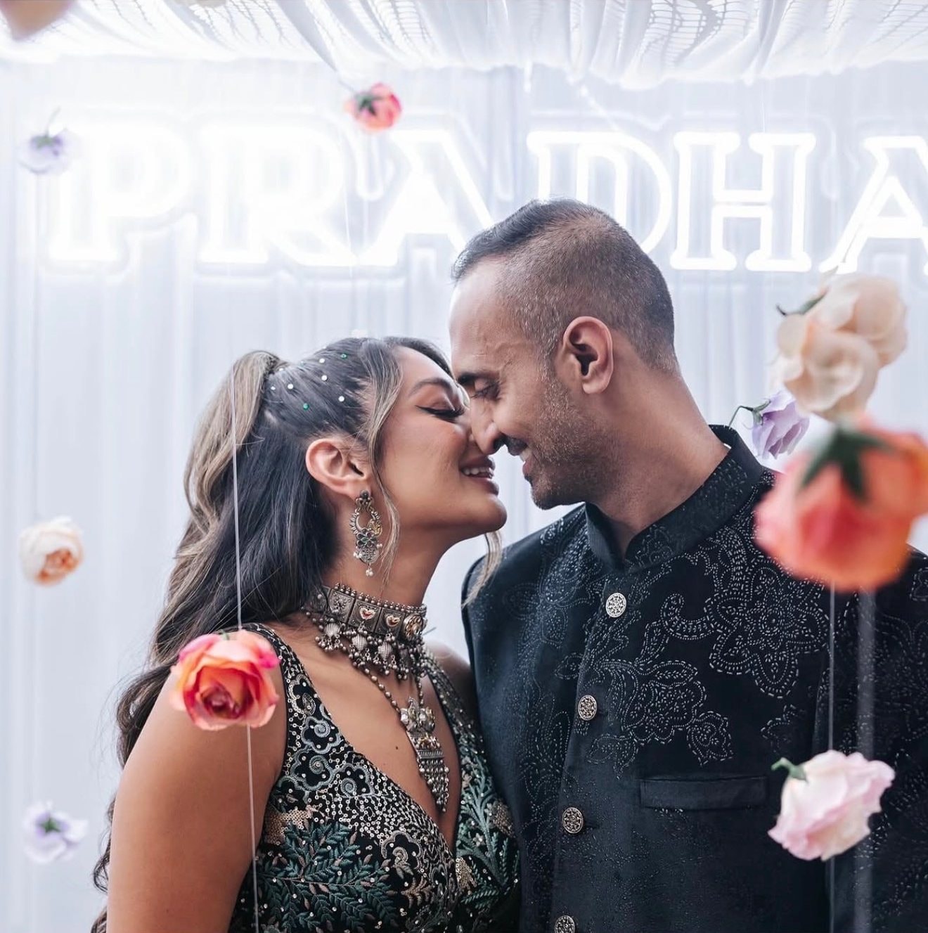 A couple leans in for a kiss in front of a glowing neon sign and hanging roses, the woman in a dark sequined outfit and the man in a black embroidered jacket.