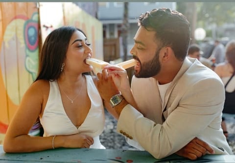 A couple in white and cream summer outfits share a popsicle from both ends at an outdoor picnic table in front of a colorful mural.