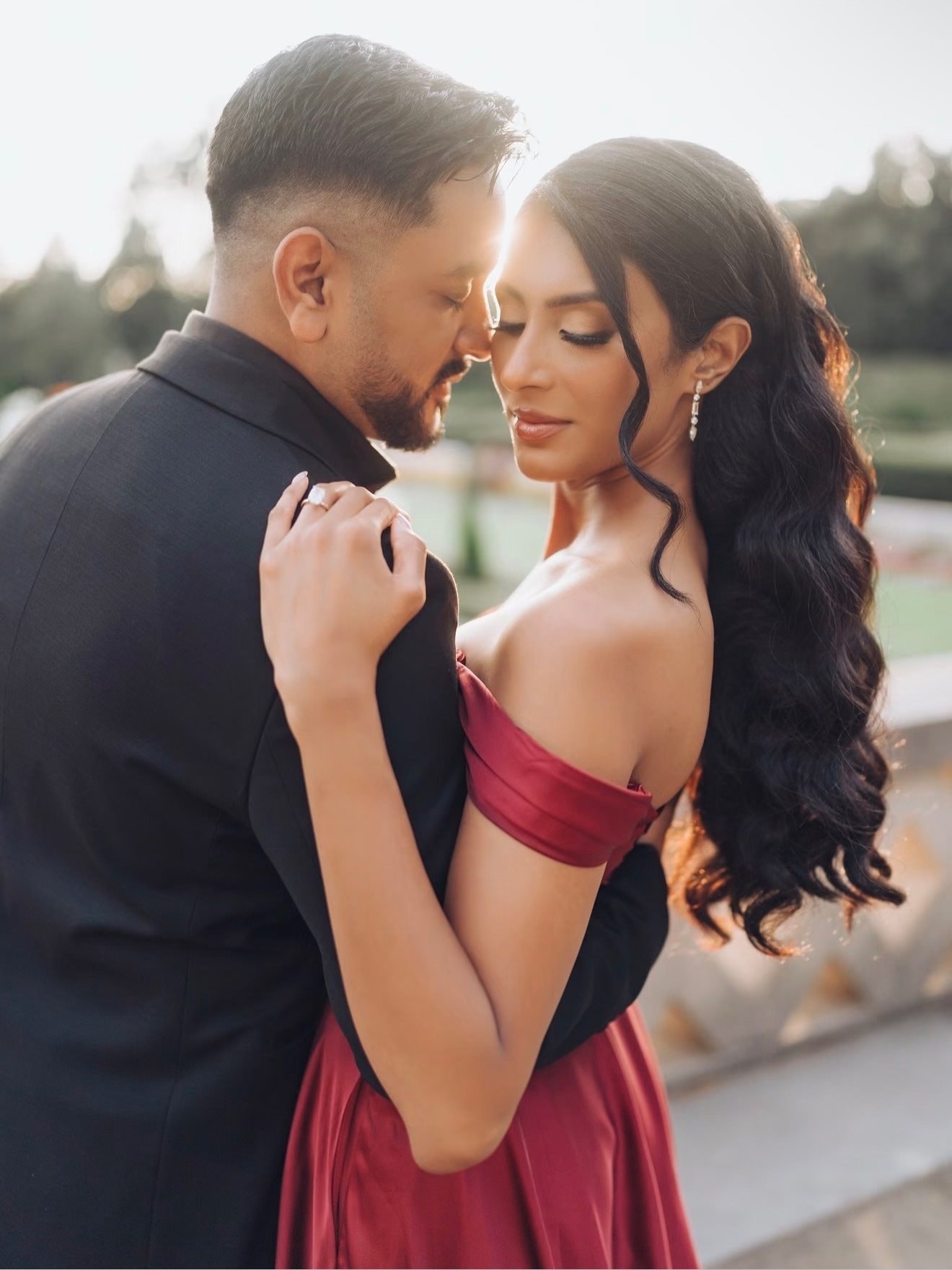 A man in a black suit holds a woman in a red off-the-shoulder gown close, her long dark curls lit by the evening sun.