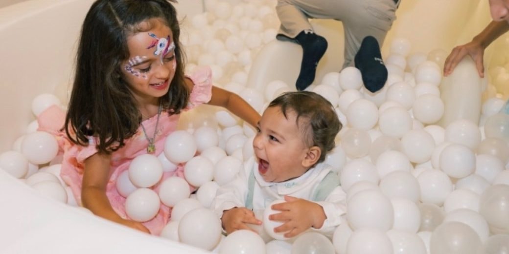 A laughing baby and a girl with a painted face play in a ball pit full of white balls.