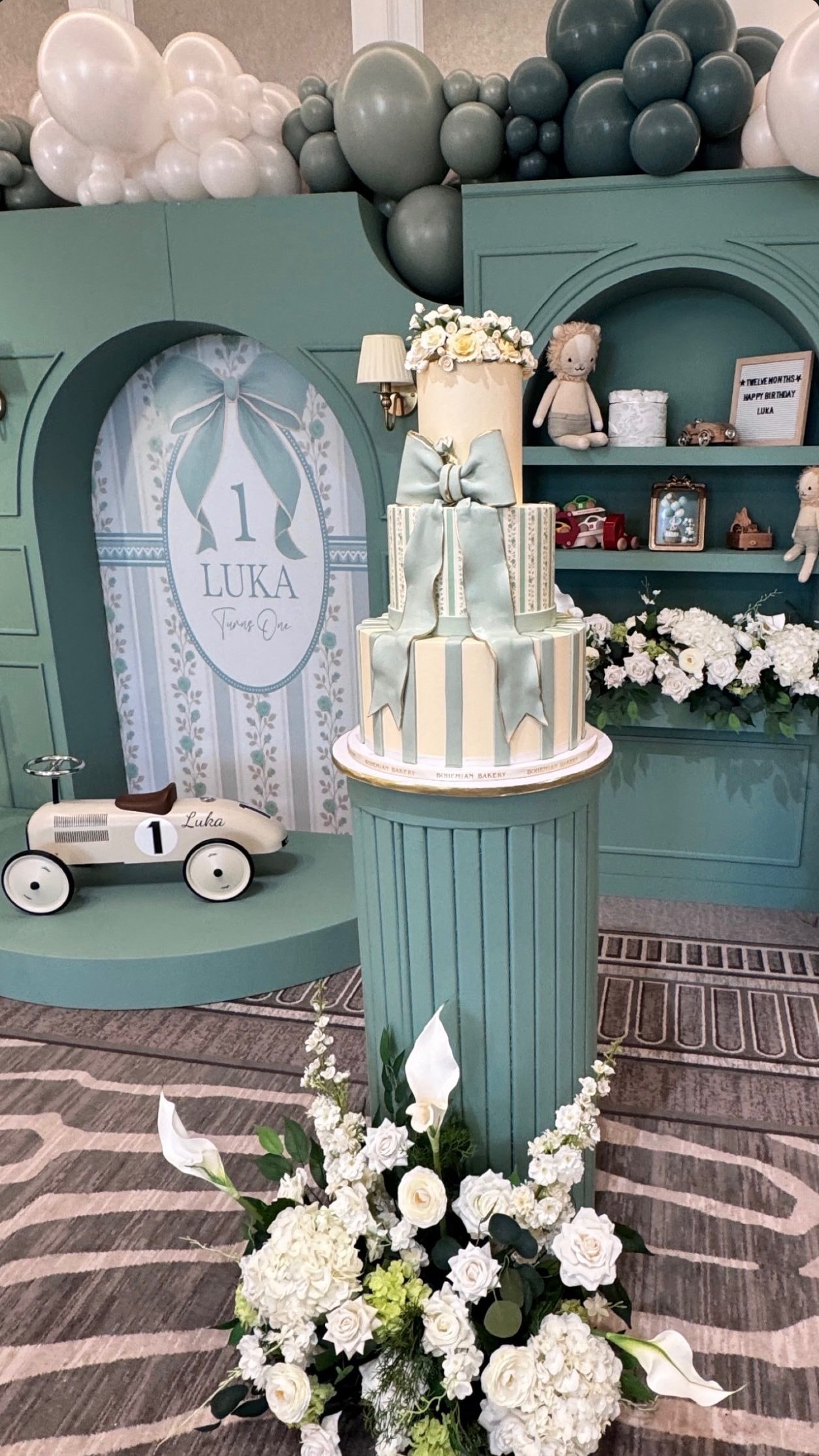 A tiered cake tied with a large sage bow on a teal fluted pedestal, in front of a teal backdrop with arched shelves, a toy race car and white flowers.