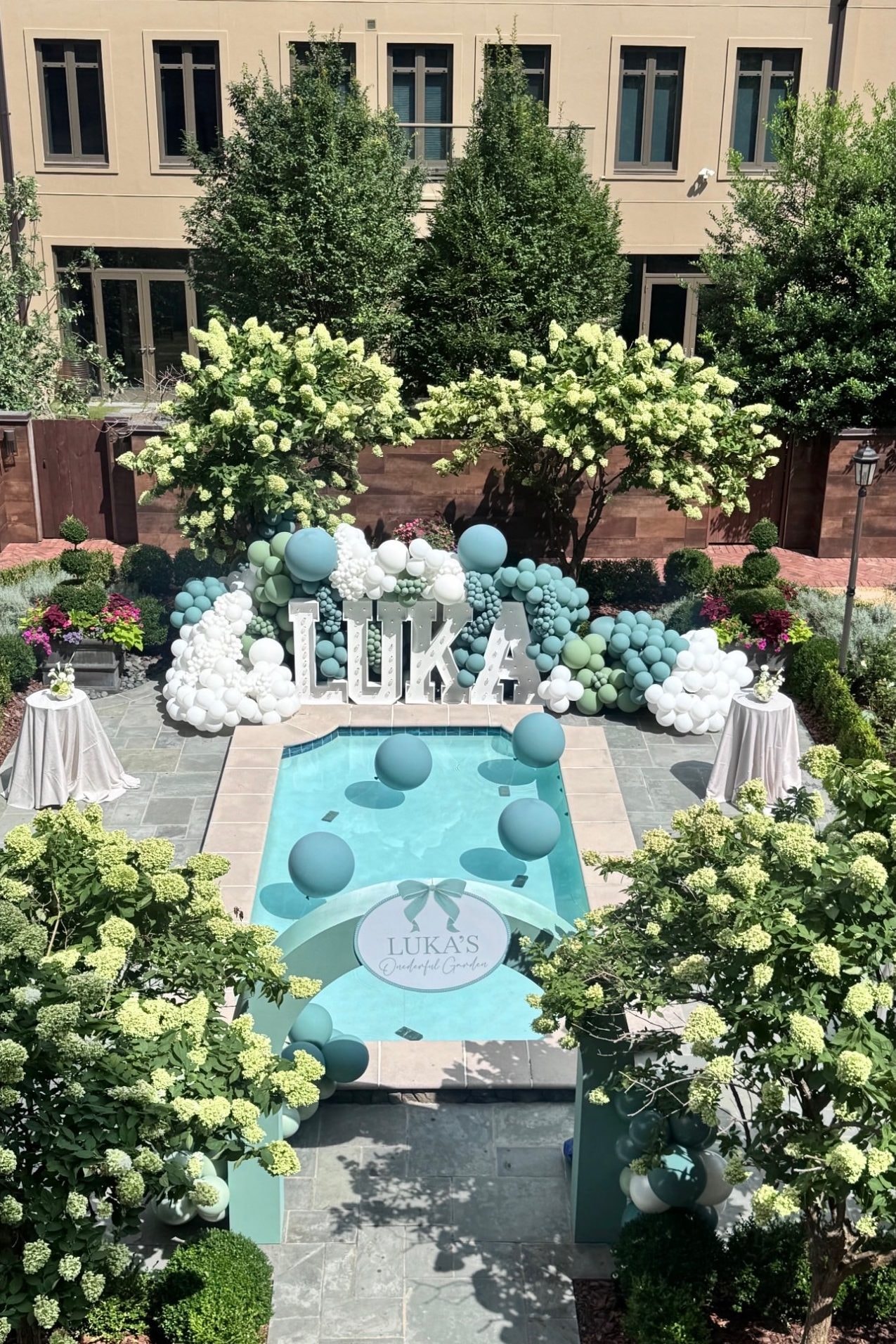 A courtyard pool dressed for a first birthday, with marquee letters spelling the birthday child’s name, teal and white balloon garlands, balloons floating on the water and white hydrangeas all around.