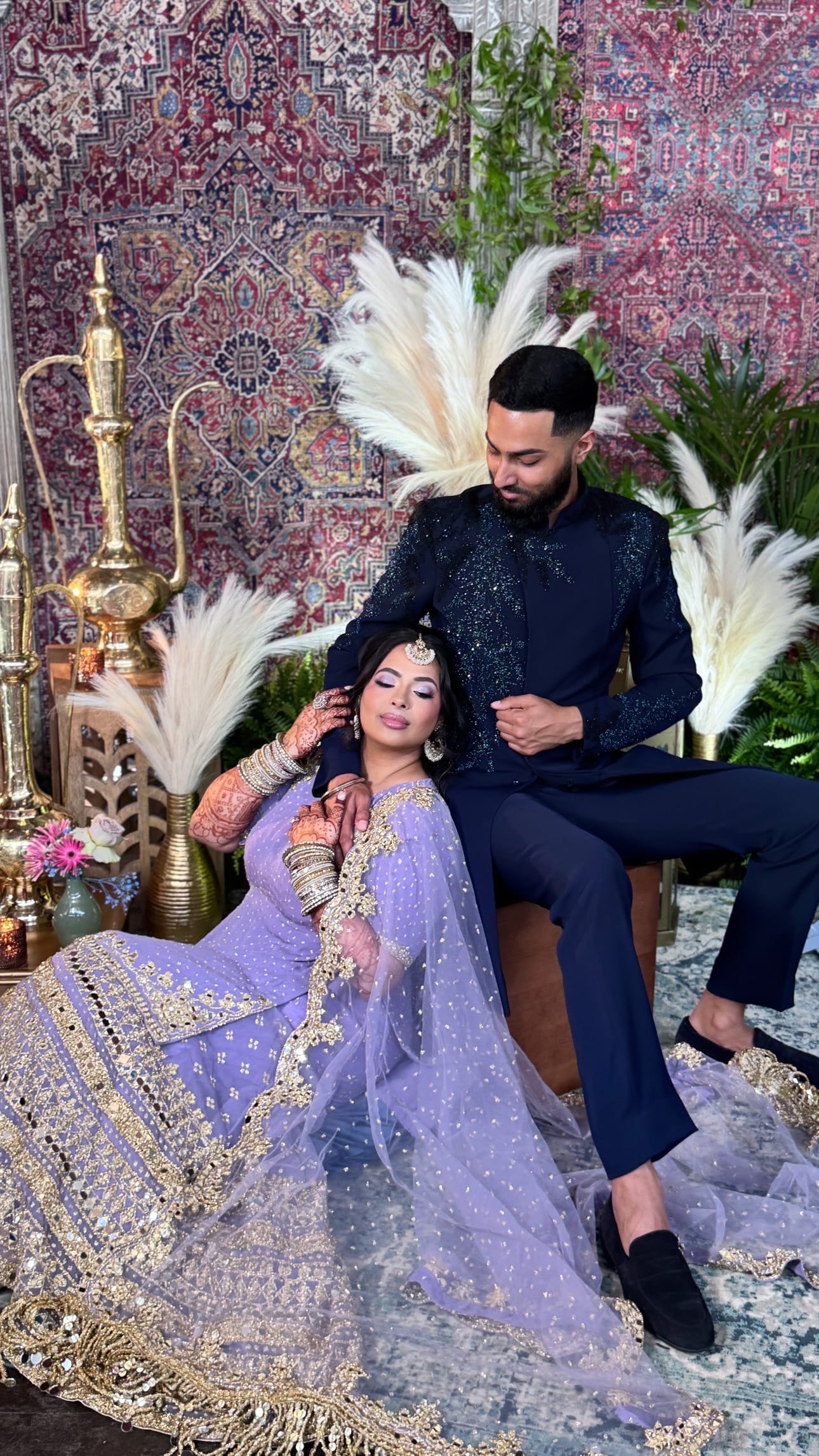 A woman in a lavender embroidered lehenga with mehndi-covered arms rests against a man in a navy beaded sherwani, seated before hanging rugs, pampas grass and tall brass lanterns.