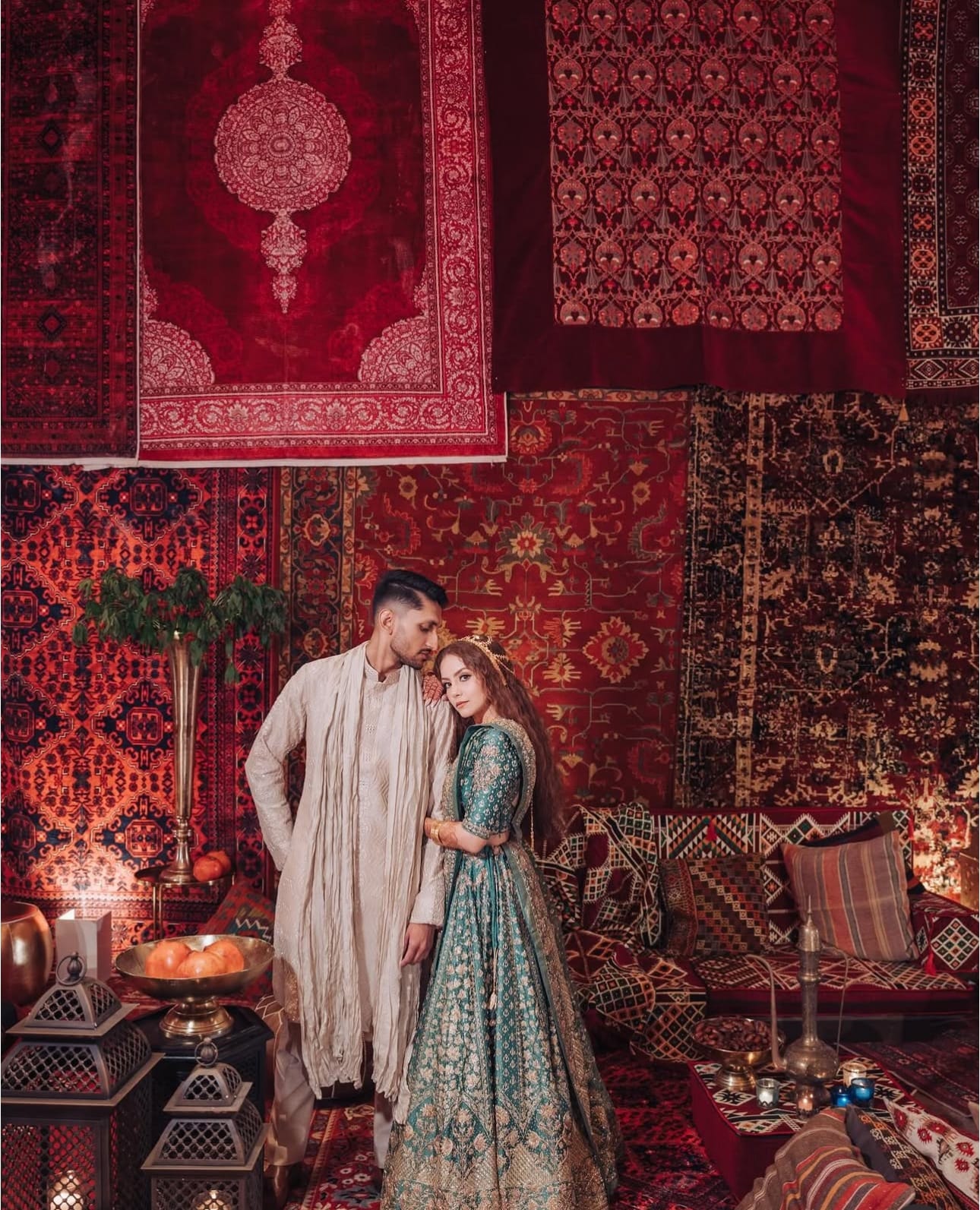 A couple stands close in a lounge walled with hanging red Persian rugs, the woman in a teal embroidered lehenga and the man in a cream sherwani, with lanterns, brass bowls of oranges and floor cushions around them.