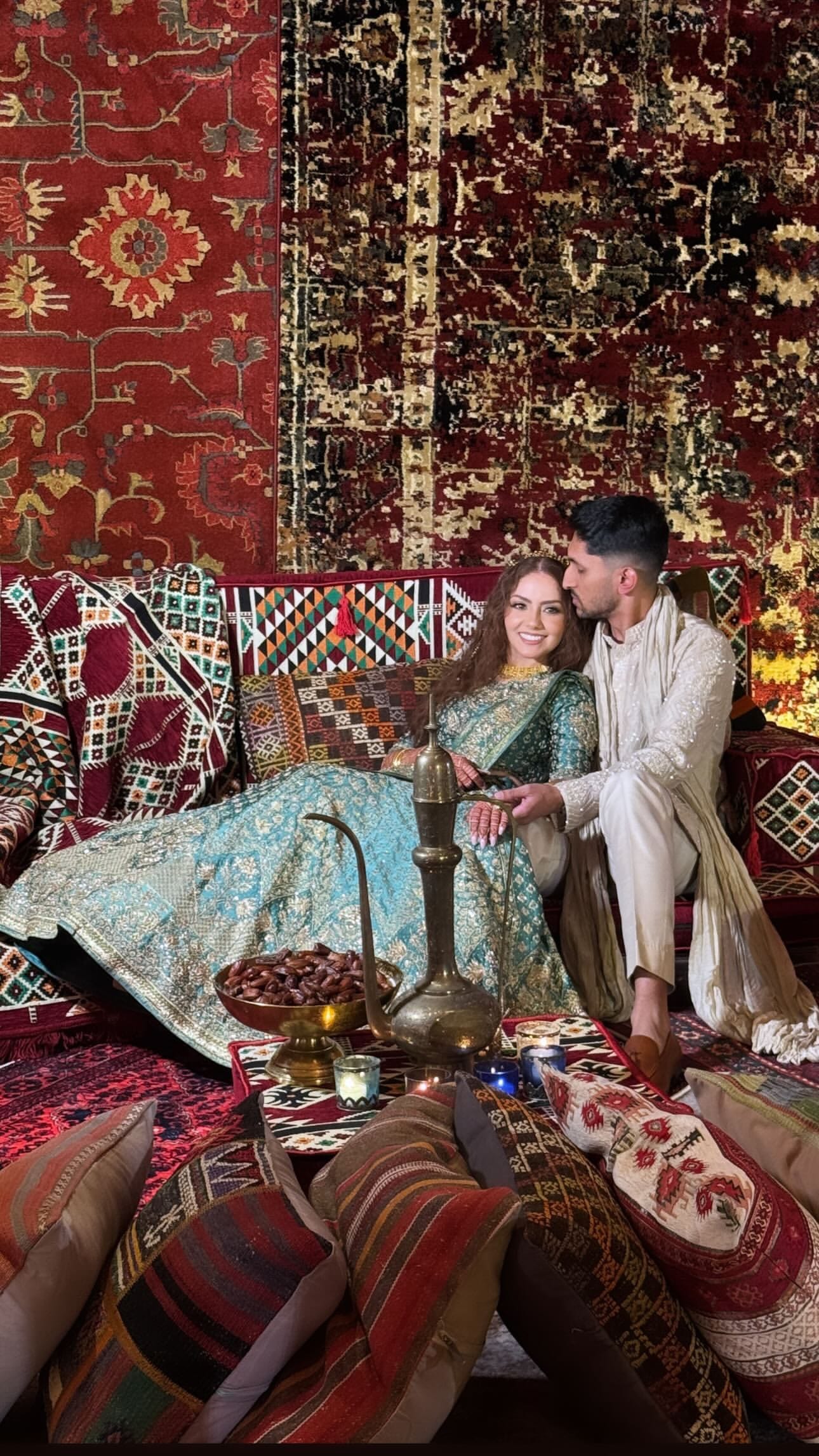 A couple reclines on a low lounge draped in layered red and gold rugs and kilim cushions, the woman in a turquoise sequined lehenga, with a brass pitcher, dates and candles on a low table.