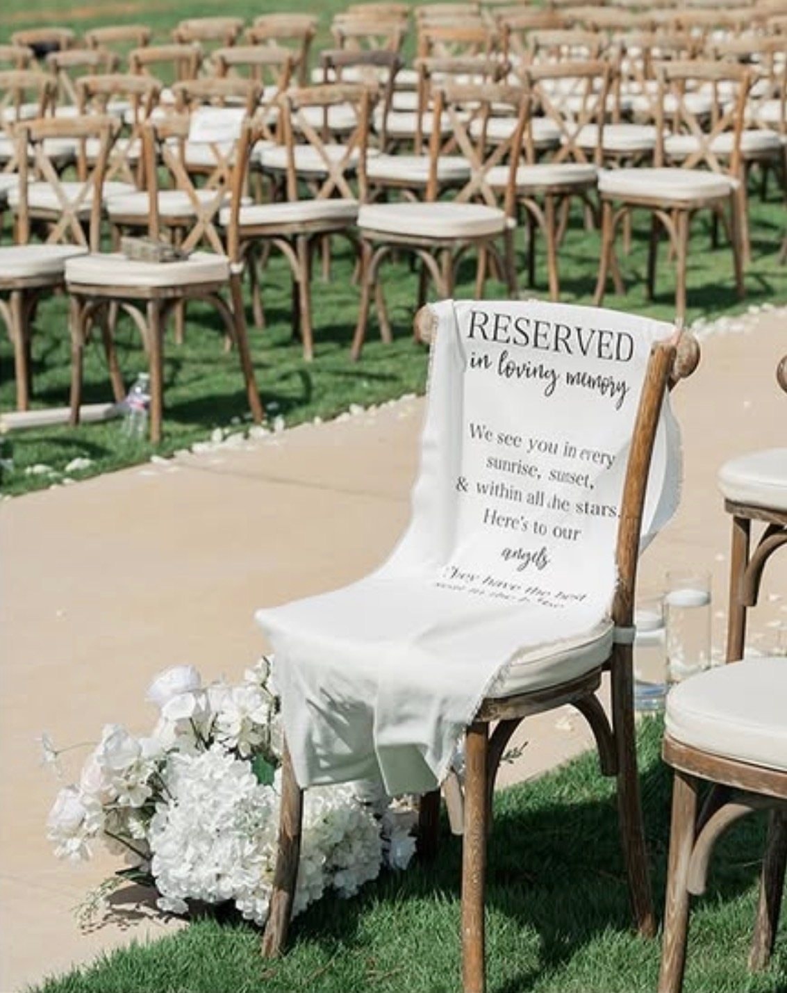 A wooden ceremony chair at the end of the aisle draped with a white sign reading ‘Reserved in loving memory’, with white hydrangeas at its feet.